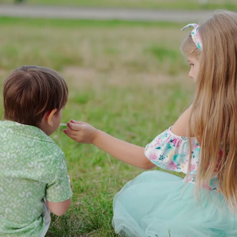 backside view of a baby boy and a girl sitting on grass. little sister feeding her smaller brother with a spoon outdoors.