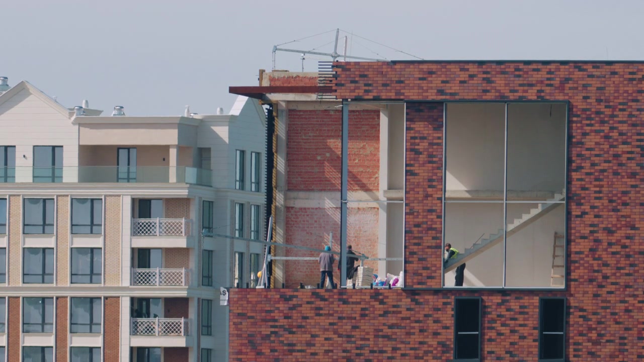 Lavoratori all'interno di un moderno edificio in costruzione, con pareti in mattoni a vista, interni grezzi e ampie aperture finestrate.
