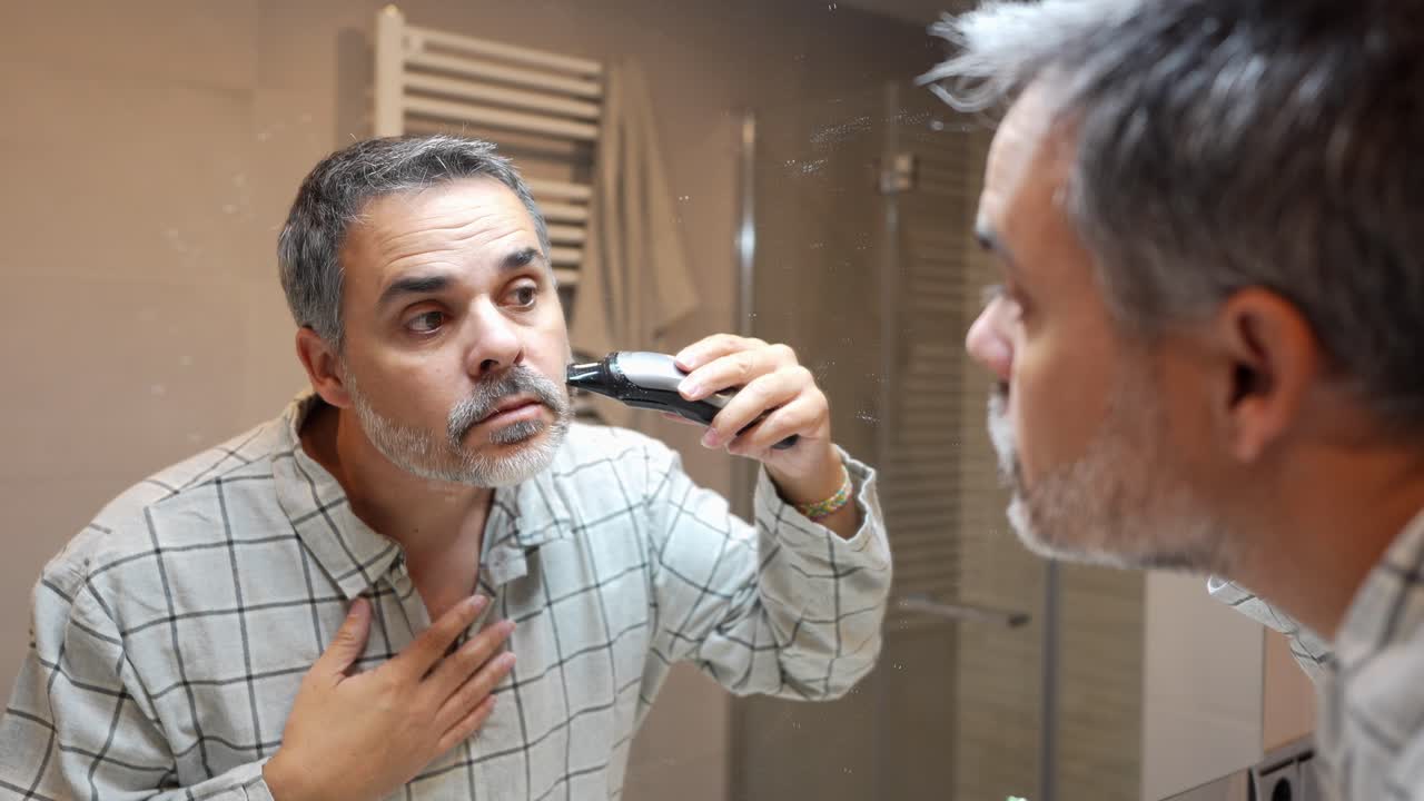 Man Trimming Beard with Electric Shaver in Bathroom Mirror