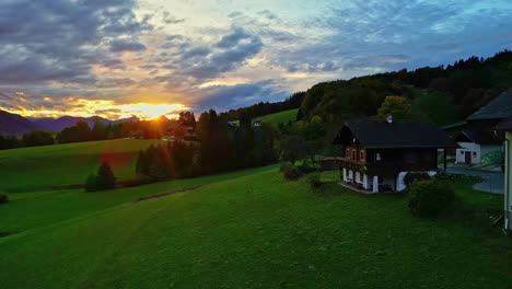 flying along the green landscape near houses and trees with sun setting behind the mountain summits in the far distance