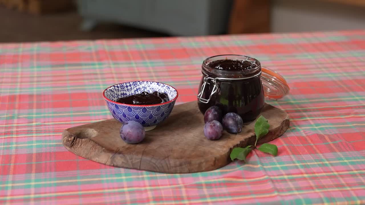 A jar and a decorative bowl of thick, dark homemade plum jam are presented on a rustic wooden board, accompanied by fresh plums and a twig with leaves