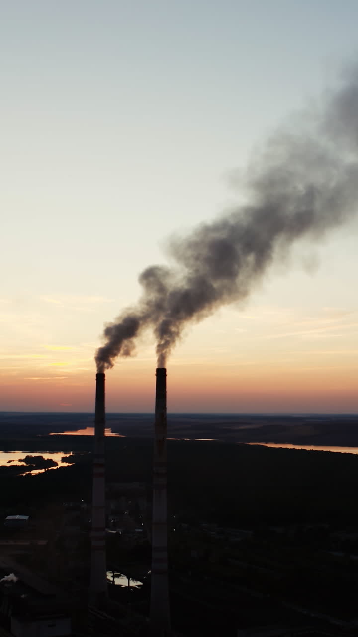 Ecological disaster. Dirty emissions from industrial pipes on the natural environment in the evening. Factory near the river at sunset. Vertical video