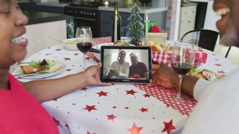 una pareja afroamericana sonriente usando una tableta para una videollamada de navidad con la familia en la pantalla