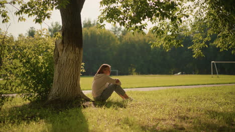 woman sitting thoughtfully by large tree in sunlit park, sunlight casting gentle shadows around her, background includes lush green trees, distant football goalpost, and car in motion