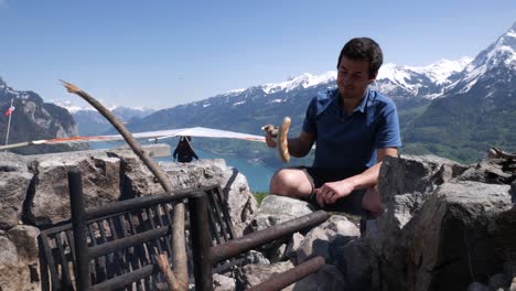 slow motion shot of a young man grilling a sausage over a bonfire while a delta sailer starts in the background over a beautiful swiss lake in the alps of switzerland