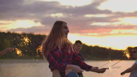 Das-Blonde-Mädchen-Im-Rotkarierten-Hemd-Und-Den-Jeansshorts-Tanzt-Mit-Großen-Bengalischen-Lichtern-In-Den-Händen-Mit-Ihren-Freunden-Am-Strand.-Dies-Ist-Ein-Wunderschöner-Sommerabend-Am-Seeufer-In-Der-Nähe-Des-Waldes.