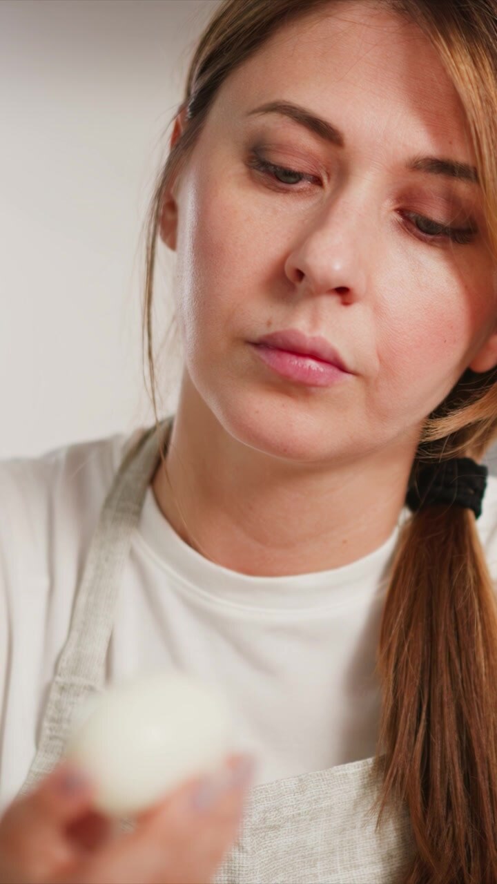 Woman holds and examines white egg showcasing its smooth texture. Woman wears light colored apron over plain shirt against neutral background with soft lighting that highlights subject