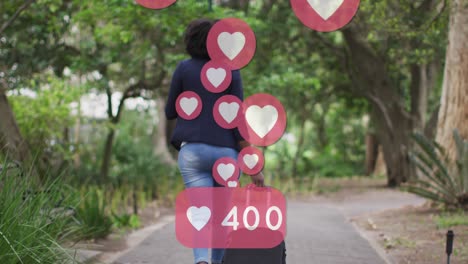 Animation-of-social-media-heart-icons-over-african-american-woman-riding-bicycle-in-park