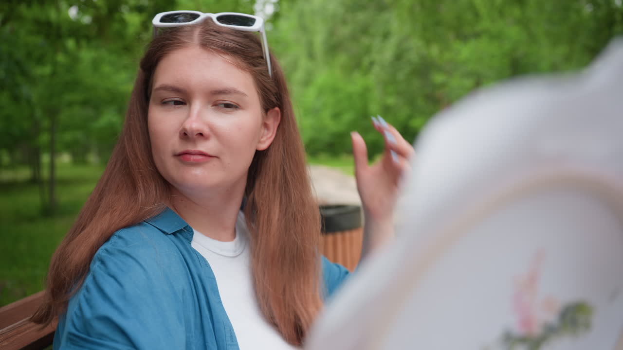Woman sitting on park bench gazing at embroidery work held in hand as if studying reflection, surrounded by lush forest trees, warm daylight creating peaceful thoughtful mood