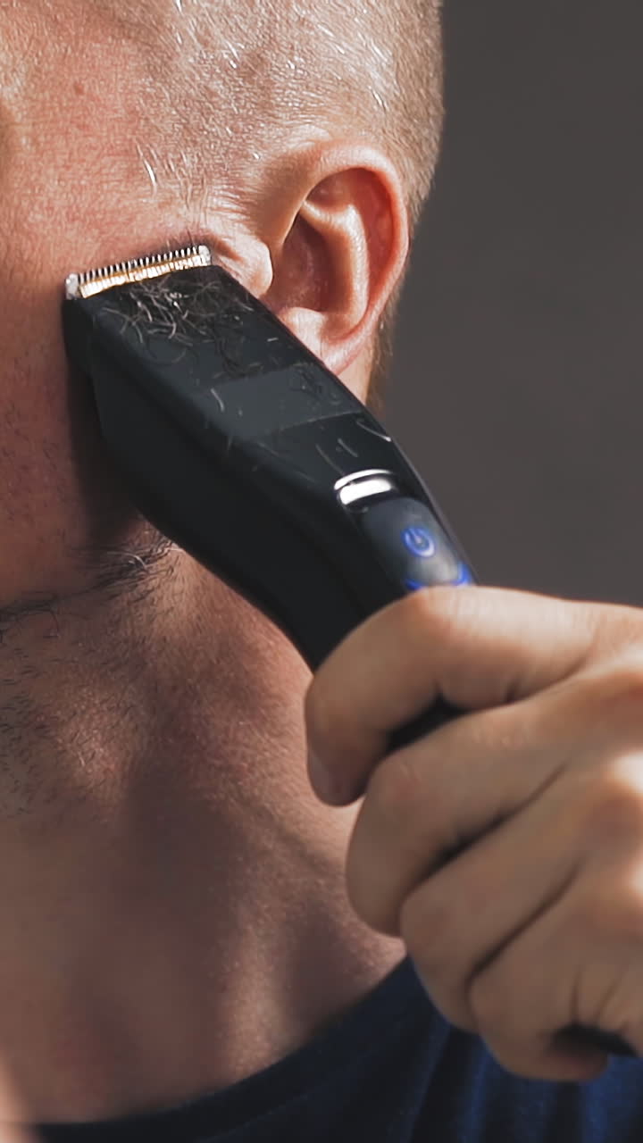CU shot, slow motion: young bald guy in black t-shirt shaves beard with modern electrical trimmer looking in large mirror in bathroom closeup