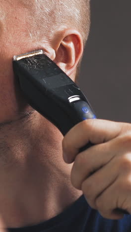 cu shot, slow motion: young bald guy in black t-shirt shaves beard with modern electrical trimmer looking in large mirror in bathroom closeup