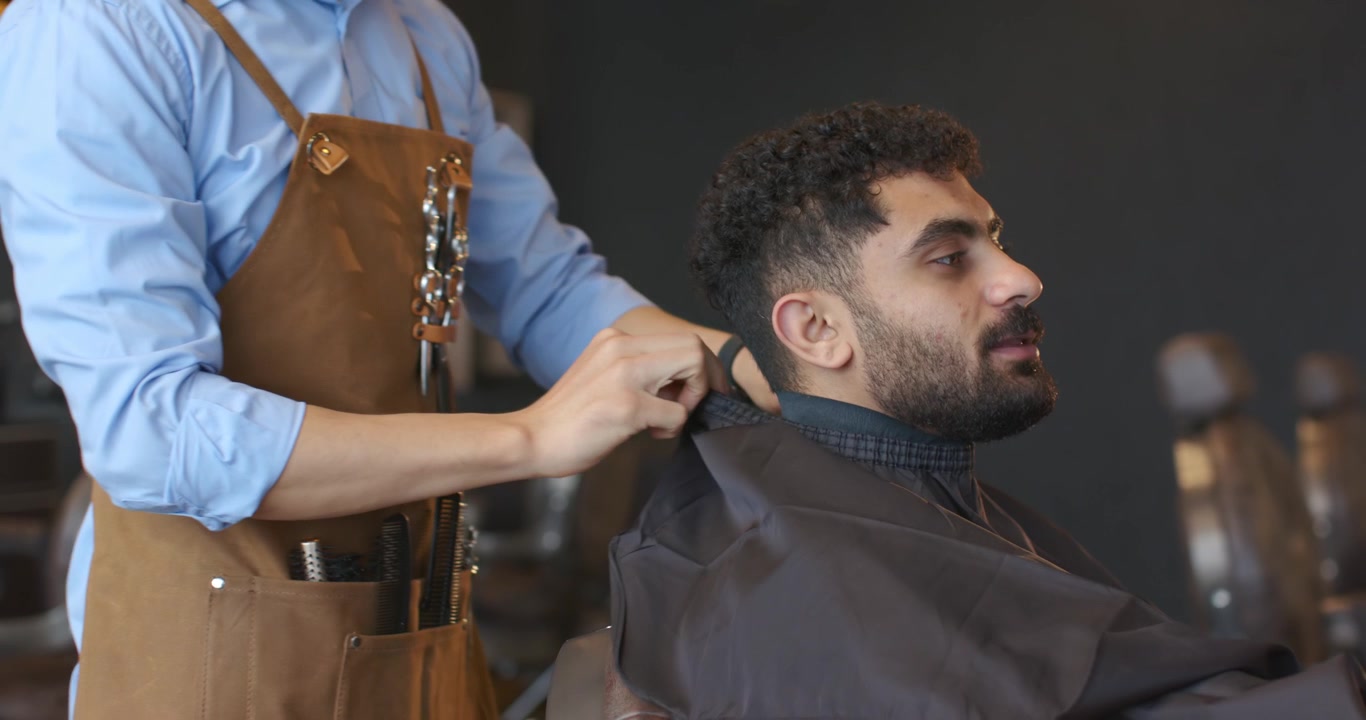 Barbiere con grembiule che avvolge la mantella al collo del cliente in barberia, fissando la chiusura per il taglio di capelli