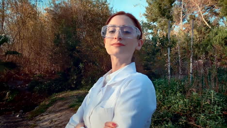 una joven científica caucásica en un arroyo, con gafas protectoras y una bata de laboratorio, cruzando los brazos y sonriendo con confianza a la cámara