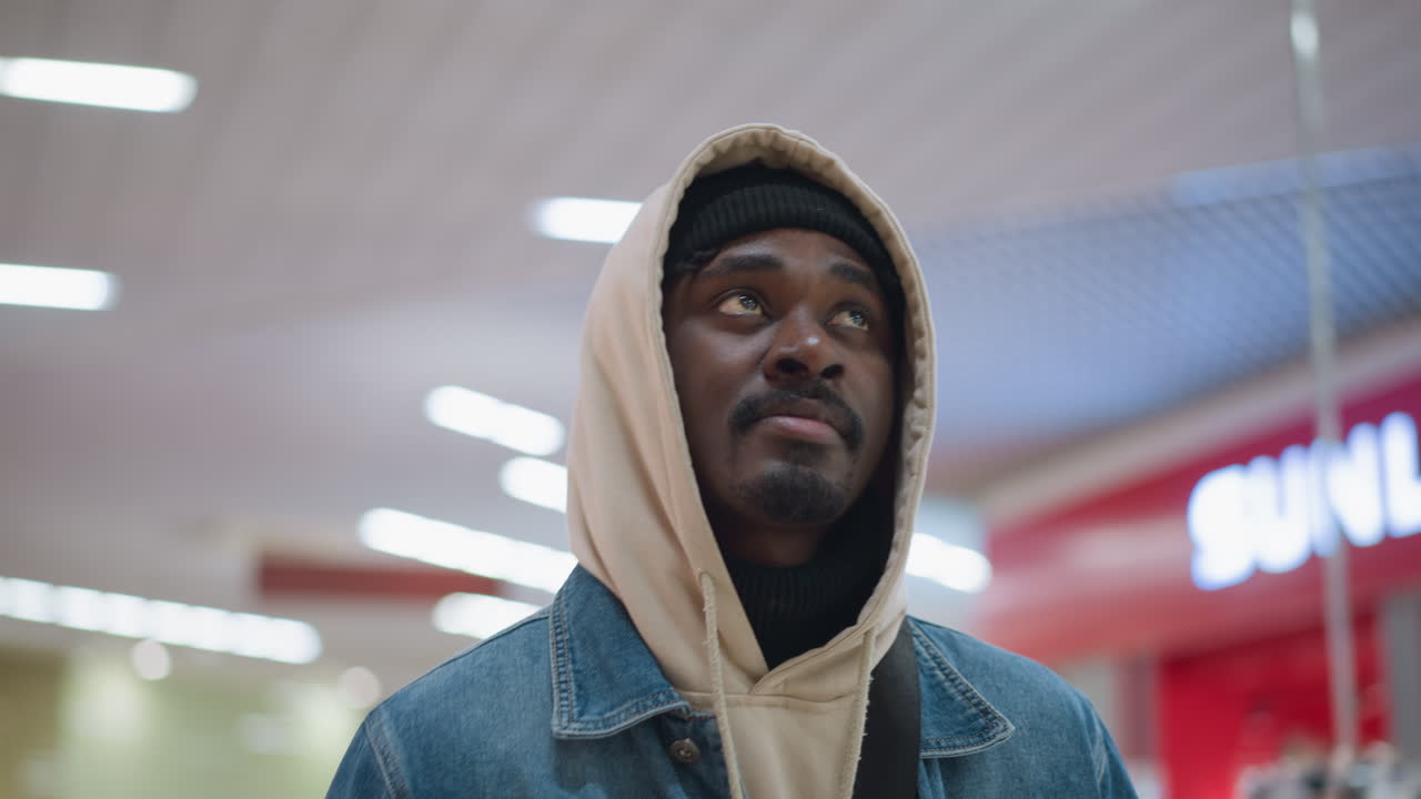 young male student wearing hoodie and denim jacket looks thoughtfully sideways standing in bright indoor campus hallway with modern ceiling lights backpack strap visible capturing calm moment