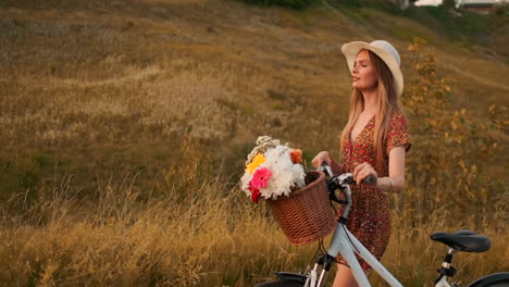 slow motion: young sexy smiling blonde woman in hat and light brown dress walking with bike and flowers in basket on field in summer at sunset. moves in the direction of the camera.