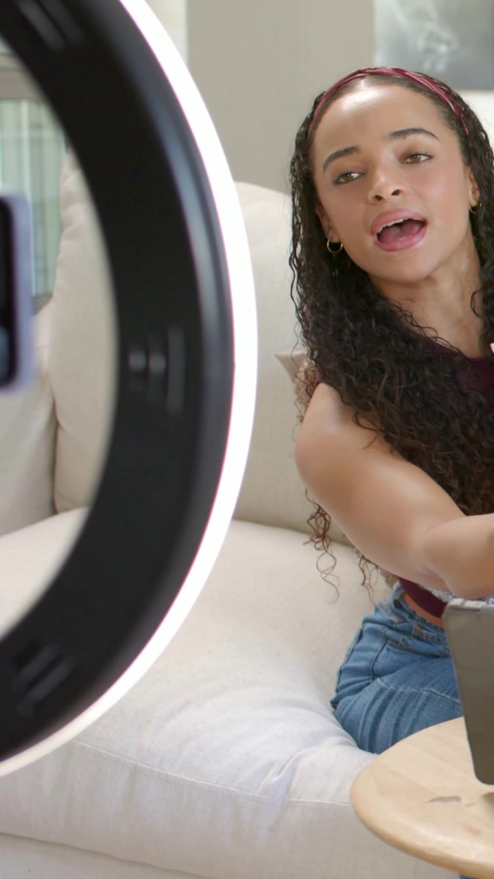 Vertical video: Ring light turning on, woman leaning on couch showing white product maroon top