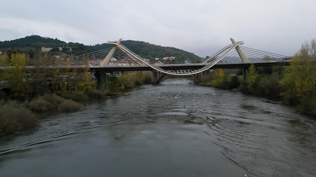 Millennium Bridge over the Mi&ntilde;o River in Ourense, Galicia, Spain, drone pullback above autumnal trees and cloudy grey sky