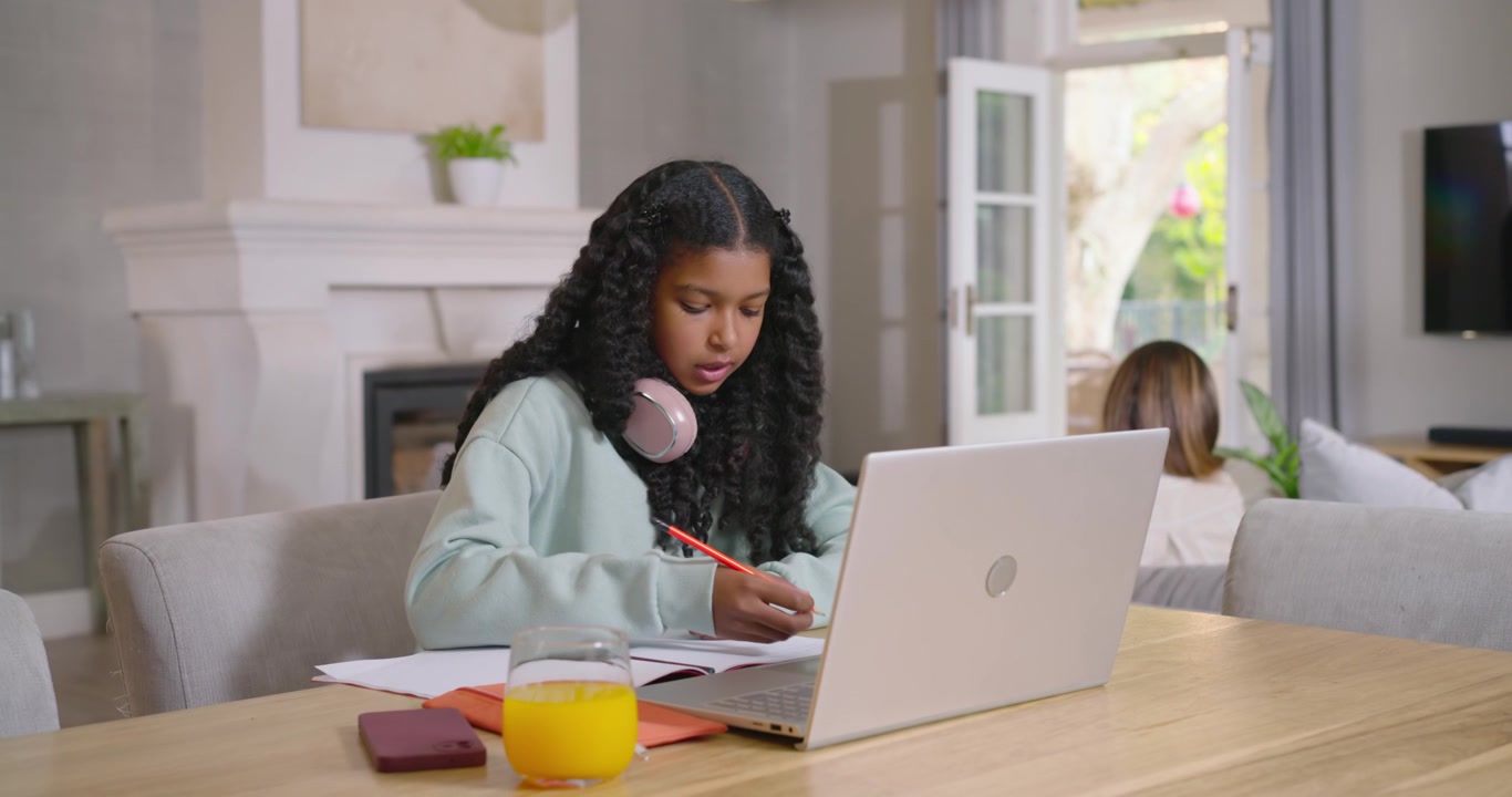 Afroamerikansk mor og tenåringsdatter, der tenåringen skriver med en oransje blyant i en laptop-klasse hjemme.