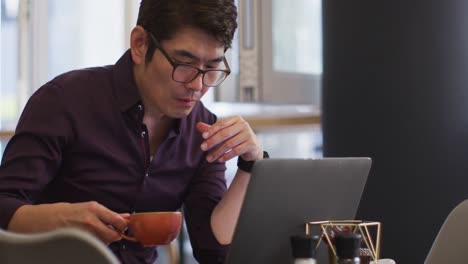 Hombre-Asiático-Con-Una-Computadora-Portátil-Tomando-Café-Mientras-Está-Sentado-En-Una-Cafetería