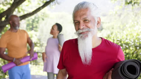 portrait of happy biracial man with white beard and yoga mat out in sun with friends, slow motion