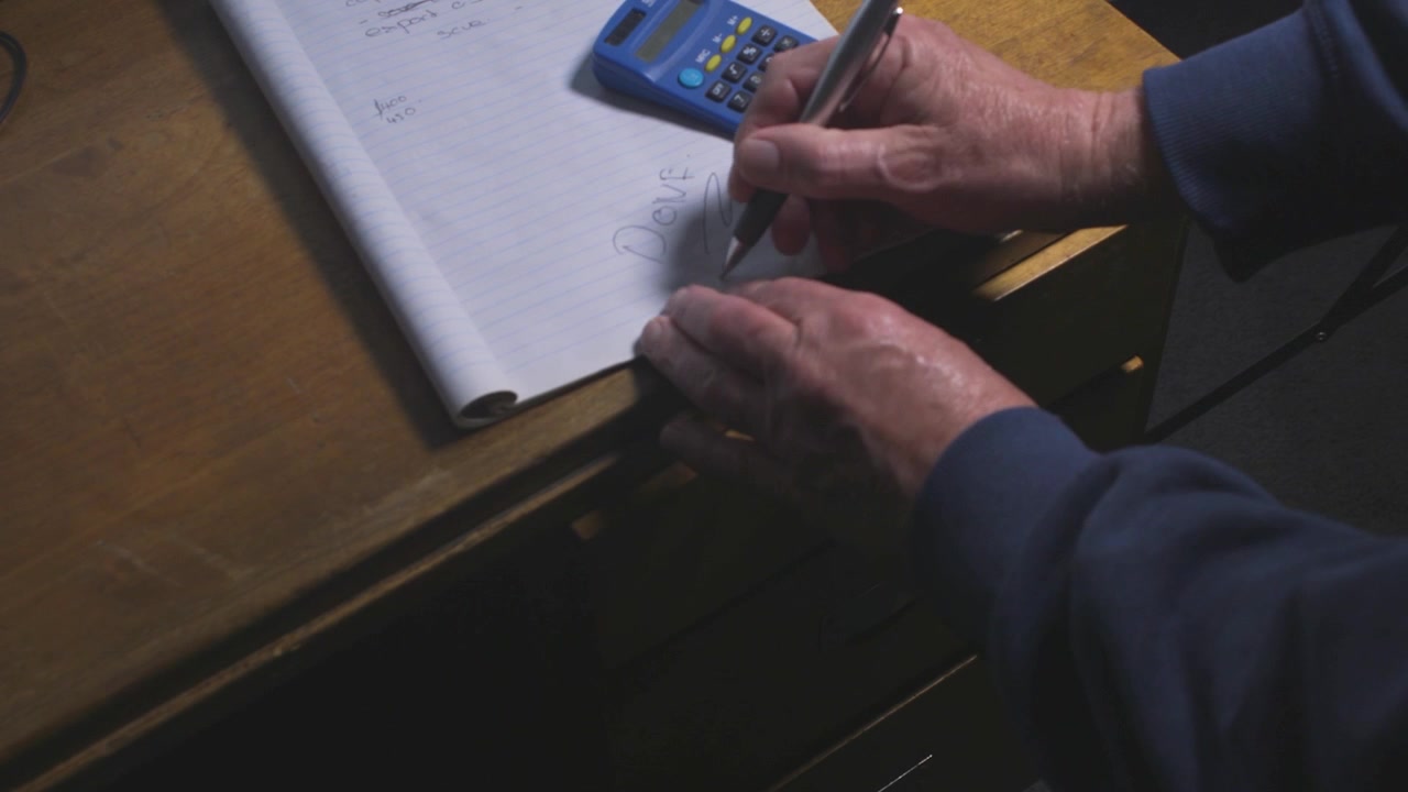 Male worker opens a filing drawer, picks up a pen beside a blue calculator, and writes notes on a desk pad.