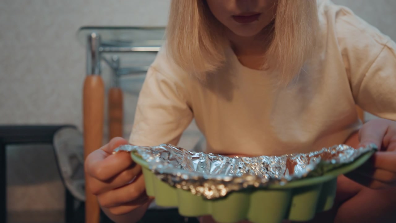 Piernas en ángulo bajo junto a la rejilla del horno. Un panadero casero arrodillado para deslizar una bandeja de silicona forrada con papel de aluminio en el horno. Destellos cálidos de bokeh, suelo de madera, un taburete de cocina informal, tensa anticipación.