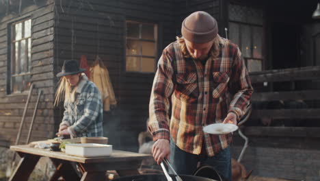 hombre cocinando carne en la parrilla mientras la esposa corta verduras