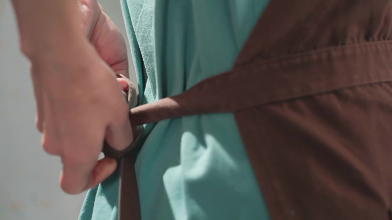 Caucasian potter tying apron strap workshop, closeup hands fastening brown apron over teal dress, warm sunlight filtering through window, calm focused preparation before clay work, textured fabric