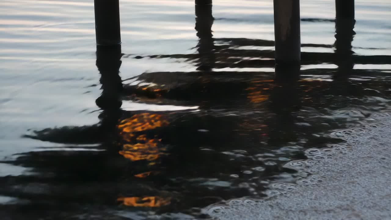 Onda lambendo a costa, causando um reflexo dourado que se move sob estacas de madeira ao anoitecer, com espuma.