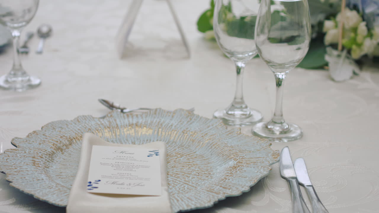 A detailed clip of an elegantly decorated wedding table. It features stemware, a white tablecloth, plates, cutlery, cloth napkins, a delicate floral arrangement, and a personalized invitation.