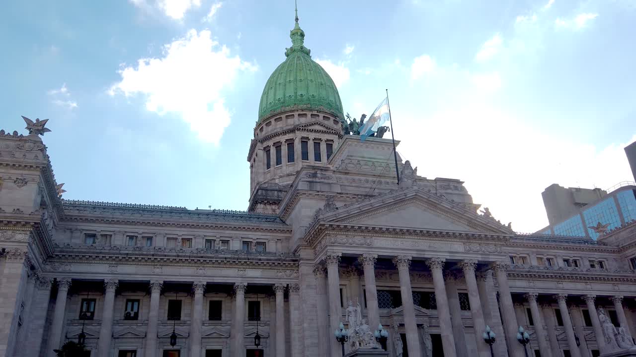 Plano em câmera lenta de baixo ângulo do Congresso Nacional da Argentina em Buenos Aires. A majestosa arquitetura neoclássica e a cúpula verde destacam-se contra um céu azul brilhante com nuvens.