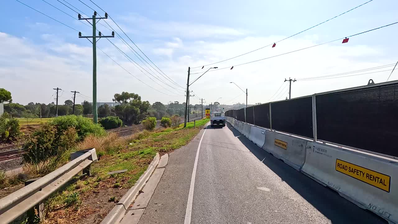 A car travels along a sunlit road in Geelong, Victoria, with clear skies and surrounding greenery
