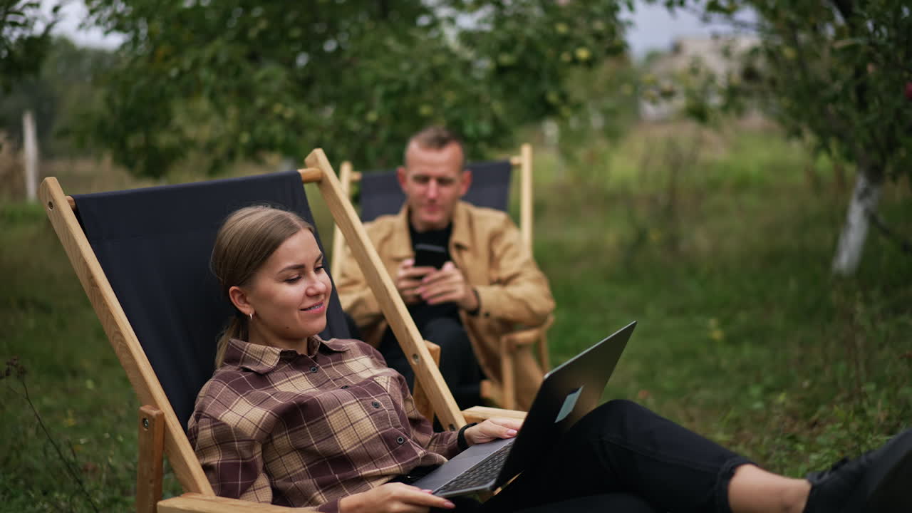 Business lady typing on laptop sitting in the garden and then leans back in chair. Busy worried man sitting beside looks at his phone.
