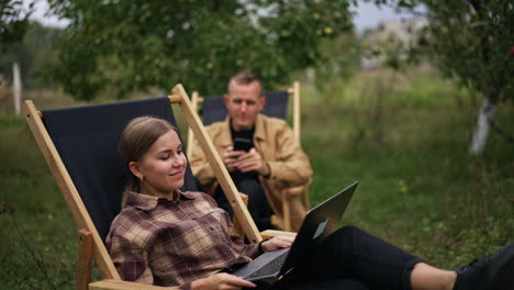 business lady typing on laptop sitting in the garden and then leans back in chair. busy worried man sitting beside looks at his phone.