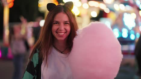 Retrato-De-Una-Niña-Feliz,-De-Cabello-Castaño-Y-Con-Un-Traje-De-Gato-Y-Una-Camiseta-Blanca,-Que-Muerde-Un-Gran-Algodón-De-Azúcar-Rosa-En-Un-Parque-De-Atracciones-Luminoso.