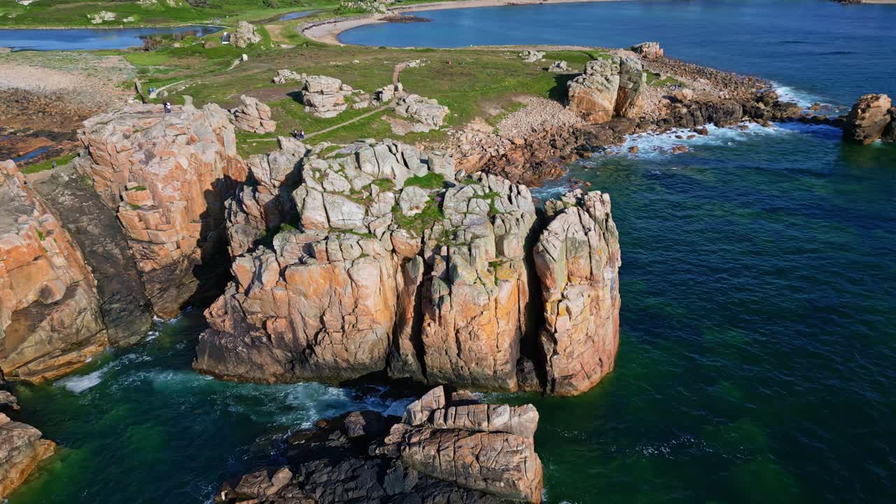 Aerial panning shot of Gouffre de Plougrescant cliffs, rock formations, and coastal trails under a clear blue sky - Brittany France