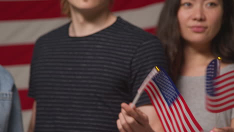 studio portrait shot of multi-cultural group of friends waving miniature stars and stripes flags in front of american flag celebrating 4th july independence day party