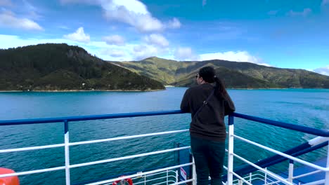 Wide-rear-view-of-asian-woman-leaning-on-railing-of-ferry-to-view-water,-mountains,-bluffs,-and-blue-sky-in-Queen-Charlotte-Sound-in-New-Zealand