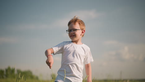 a young boy, wearing glasses and a white shirt, stands in a sunny grass field holding a piece of grass, cheerfully playing with it. the clear sky and vibrant green grass add to the joyful