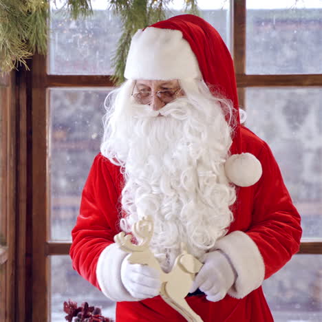 portrait of santa claus holding wooden toy deer. old santa in red costume with white beard standing near the window on christmas decorated background indoors.