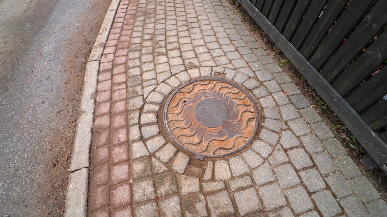 Close-up of decorative manhole cover in cobblestone sidewalk beside a curb, showing wet pavement texture and urban drainage detail after thaw.