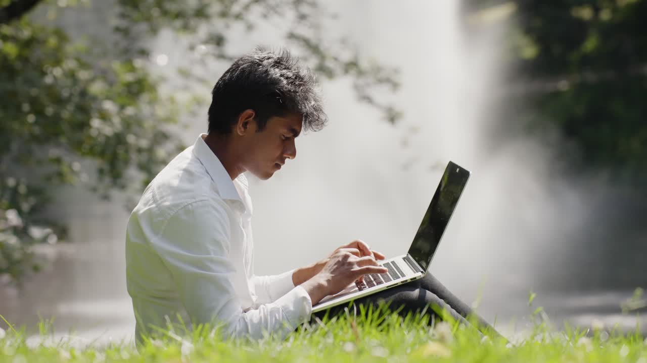 Young professional with laptop working remotely from public park, Water sprays from fountain in Background