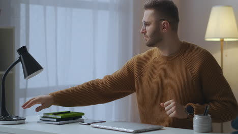 man is starting remote work with laptop at home sitting at table and opening notebook turning on table lamp typing on keyboard freelance and part-time job