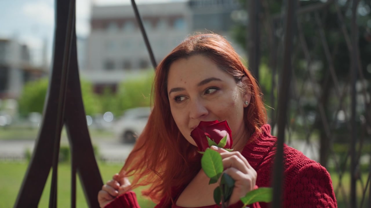 Close up of woman with red hair wearing maroon knit sweater holding and smelling single dark red rose while sitting on outdoor swing. background features blurred greenery and distant