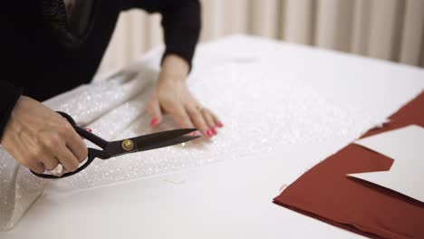 Fashion-designer-working-with-white-sparkling-fabric-at-the-studio-full-of-tailoring-tools---pattern,-scissors.-Workplace-of-seamstress.-Tailor-cuts-wedding-dress-detail-on-the-sketch-lines.-Unrecognizable-woman