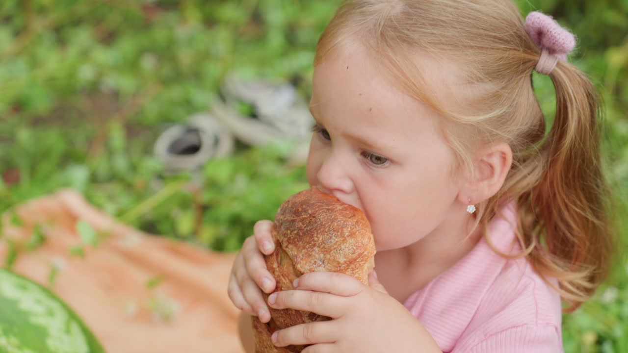 white toddler girl biting crusty loaf on orange blanket in grassy park, nibbling and inspecting crumbs, covering eye in shy moment, turning gaze toward parent, warm sunlight, candid family picnic