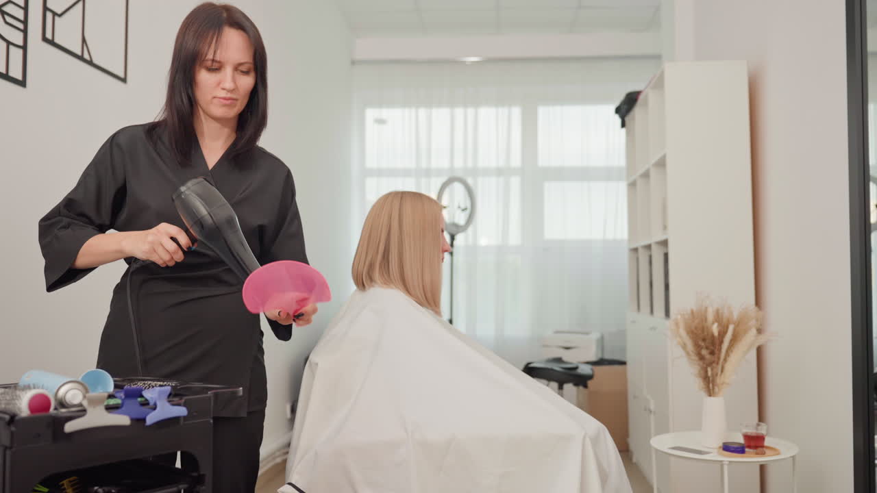 Parrucchiere che asciuga i capelli biondi con l'asciugacapelli in un luminoso salone domestico, mantellina bianca drappeggiata sulla cliente, carrello con strumenti e prodotti, luce naturale dalla finestra, delicati colpi di spazzola, arredamento neutro caldo, atmosfera rilassata