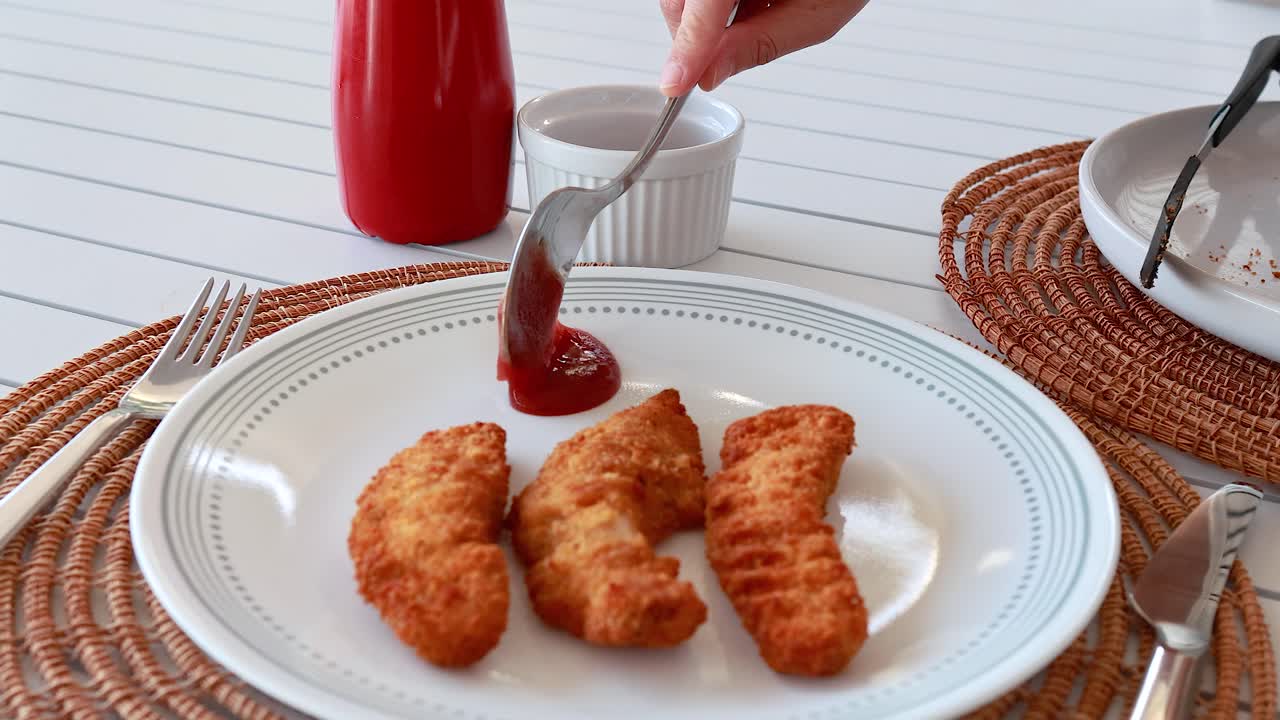 Una mano versa la salsa su striscioline di pollo fritto croccanti su un piatto bianco, posizionato su una tovaglietta intrecciata con illuminazione brillante