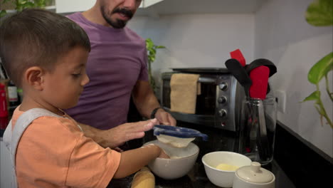 padre latino con una camiseta púrpura cocinando en la cocina con su hijo haciendo galletas tamizando harina golpeando un tamiz