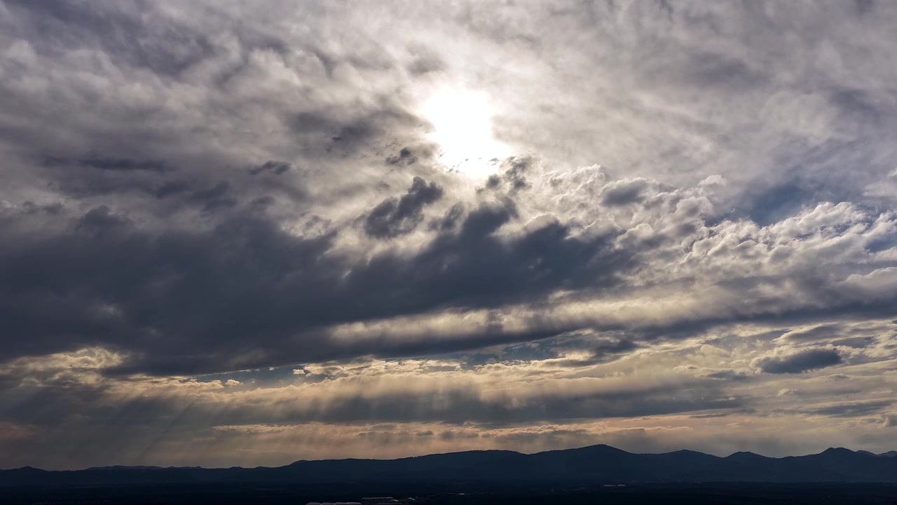 4K UHD drone timelapse of the bright sun partially hidden behind thick, dramatic cloud layers. Brilliant rays of light pierce the heavy overcast sky, rippling dynamically across the dark mountain land